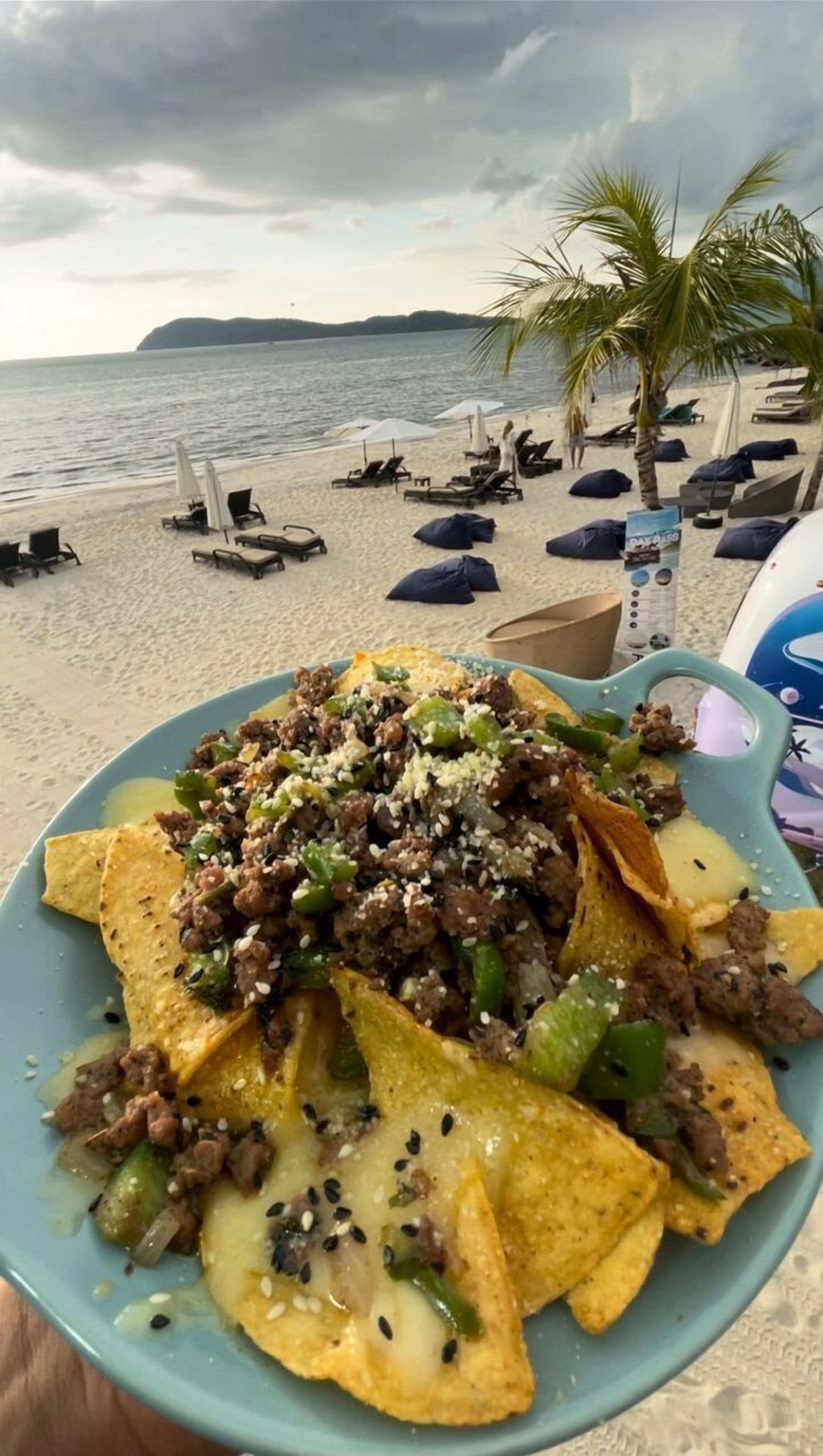 A plate of nachos with meat and cheese on a beach with palm trees and lounge chairs.