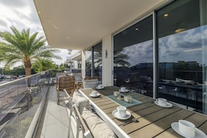 A balcony with outdoor seating, large windows, and palm trees. - Eagle Beach O condo 18 by Bocobay (Noord)