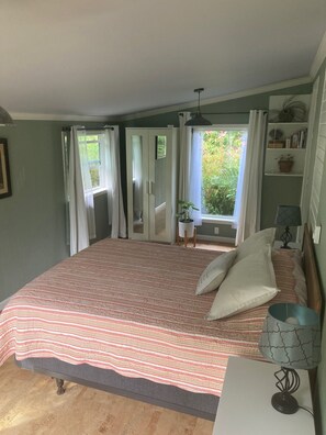A bedroom with a large bed, a bedside table with a lamp, a window with curtains, and a view of greenery outside.