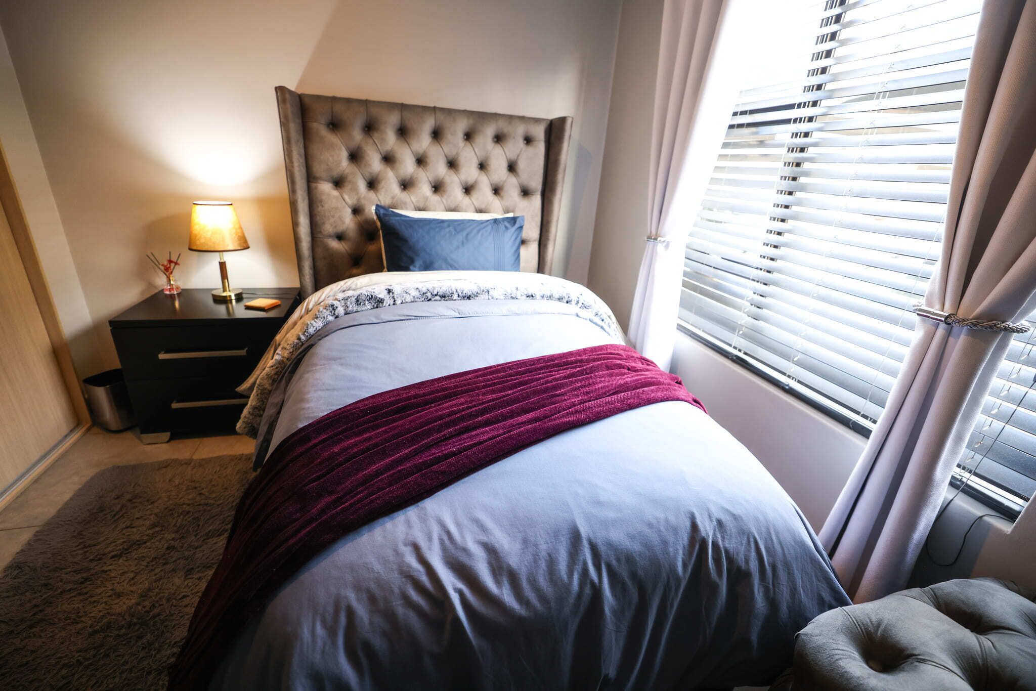 A bedroom with a tufted headboard, a bedside table with a lamp, a bed with a white and blue bedspread, and a window with blinds.