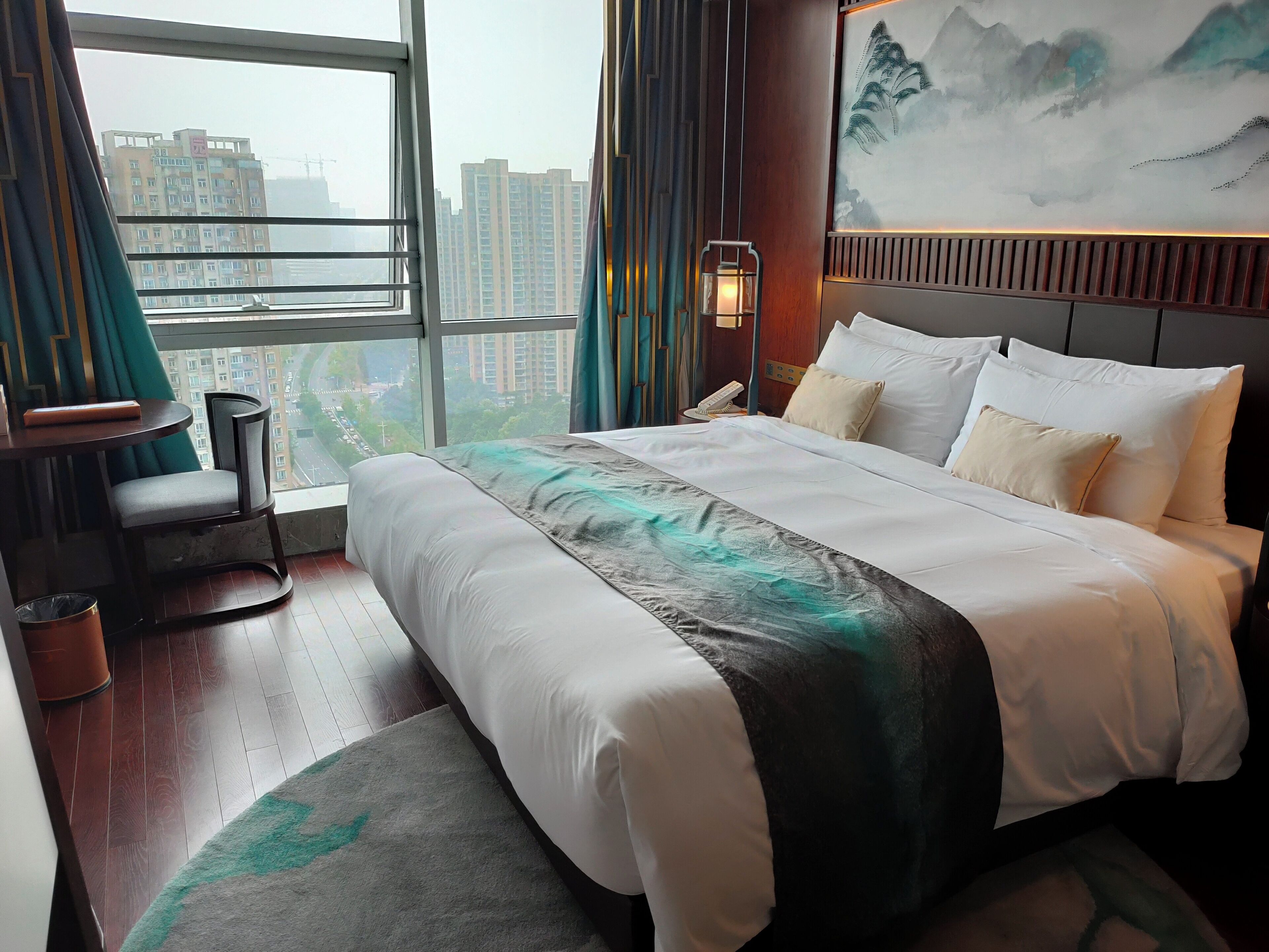 A hotel room with a large bed, a chair, a small table, a waste bin, and a view of the city through a large window.