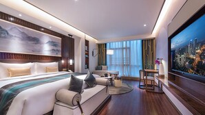 A modern hotel room with a large bed, a sofa, a dining area, and a TV displaying a cityscape.