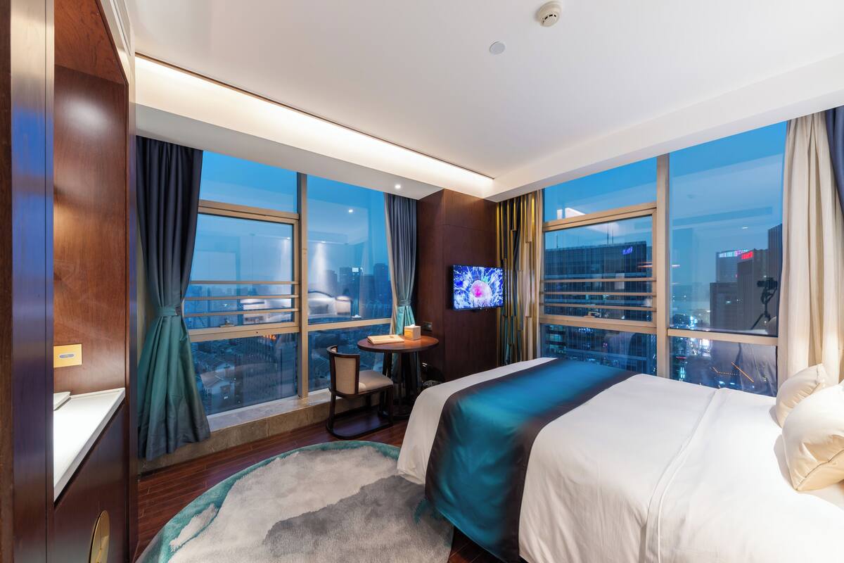 A modern hotel room with a large bed, a TV on a stand, a small table, and a view of the city through large windows.