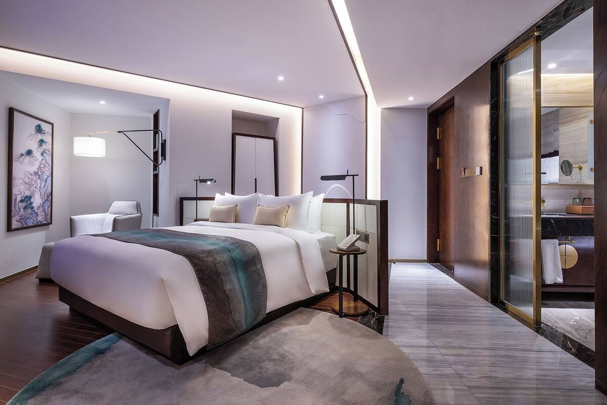 A modern hotel room with a large bed, bedside tables, a desk, and a bathroom visible through a sliding door.