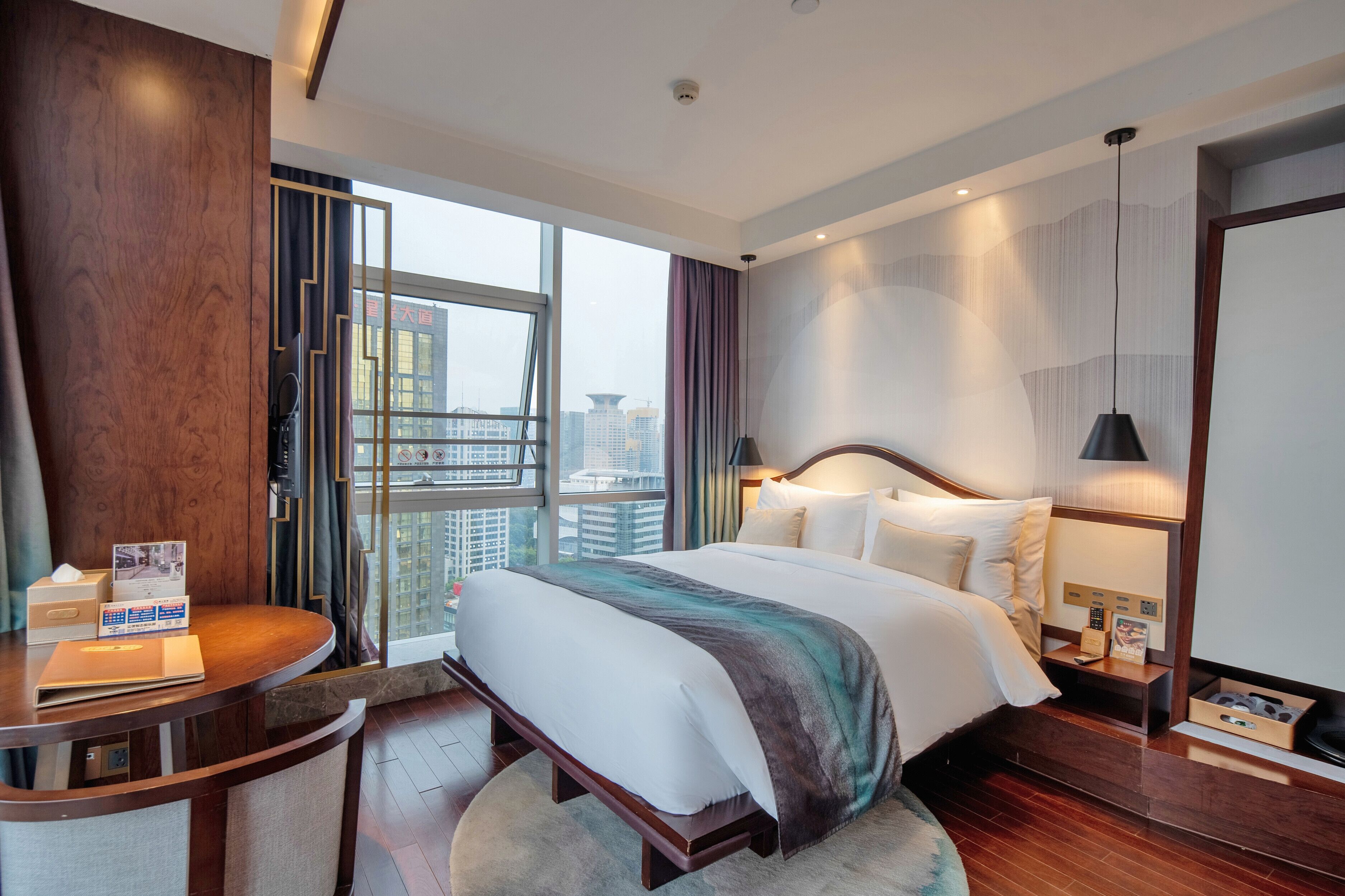 Une chambre d’hôtel avec un grand lit, un bureau, une chaise, une table de chevet et une vue sur la ville grâce à de grandes fenêtres.