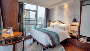 A hotel room with a large bed, a desk, a chair, a nightstand, and a view of the city through large windows.