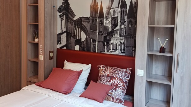 Une chambre d’hôtel avec un grand lit, des oreillers rouges et blancs, une armoire en bois et une fresque murale représentant un paysage urbain.