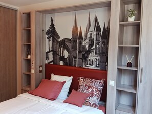 A hotel room with a large bed, red and white pillows, a wooden wardrobe, and a wall mural of a cityscape. - Studio on the banks of the Orne (Caen)