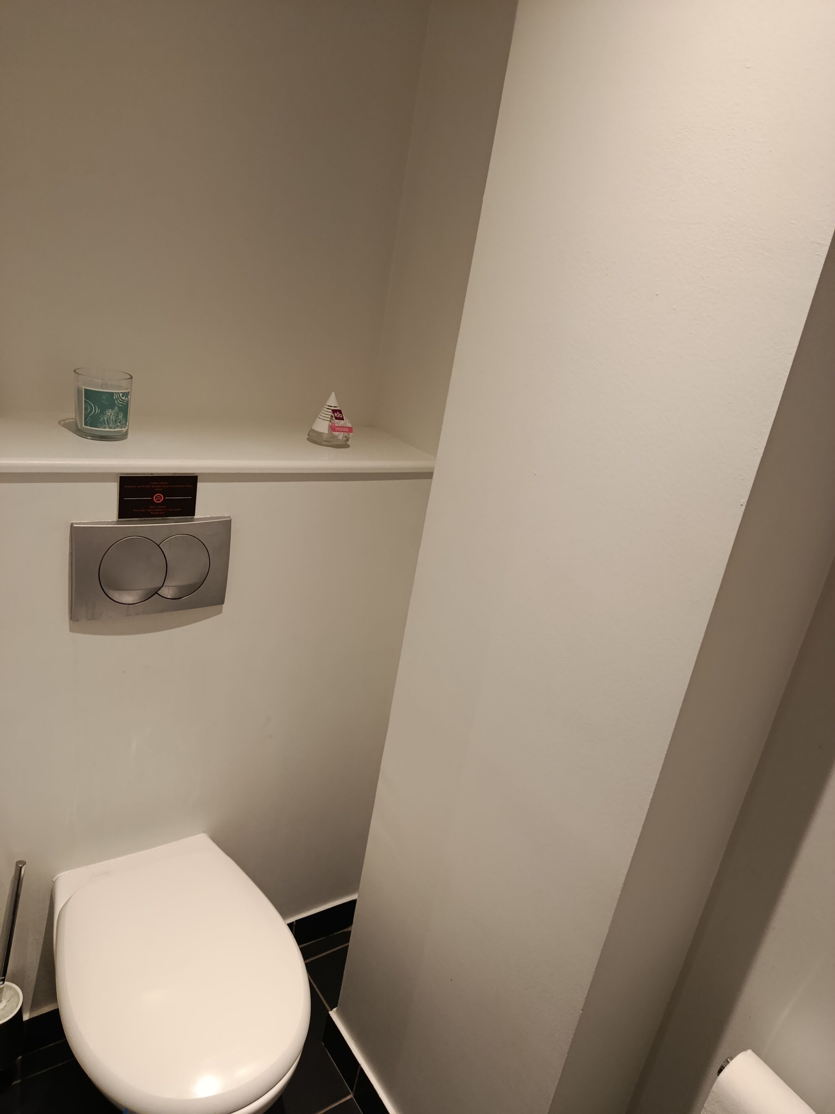 A small, narrow bathroom with a white toilet, a hand dryer, and a small shelf with a glass candle and a tissue box.