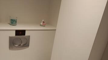 A small, narrow bathroom with a white toilet, a hand dryer, and a small shelf with a glass candle and a tissue box.