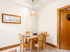 Un comedor con una mesa de madera dispuesta para cuatro personas, rodeada de sillas. Una lámpara colgante ilumina la mesa, y hay un cuadro enmarcado en la pared.