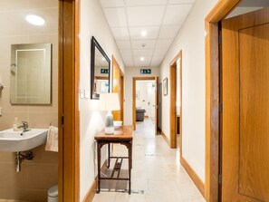 Un pasillo de hotel con un baño a la izquierda, una mesa de madera con una lámpara y un espejo.