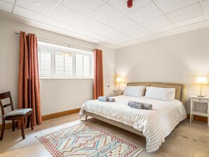 Un dormitorio con cama, dos mesitas de noche con lámparas, una silla, una ventana con cortinas y una alfombra estampada.