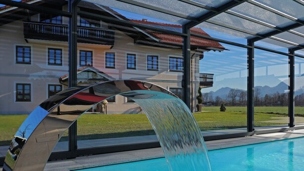 A swimming pool with a modern water feature, a glass-enclosed patio, and a large building with a red-tiled roof in the background. - Gemütliches Ferienwohnung in Feldwies mit Sauna (Ubersee)