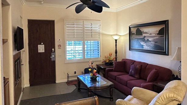 A living room with a dark ceiling fan, a red sofa, a beige armchair, a coffee table, and a painting on the wall. - Sun-Soaked Condo near Old Town (La Quinta)