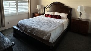 A neatly made bed with a wooden headboard, two bedside tables with lamps, a ceiling fan, and a window with blinds. - Sun-Soaked Condo near Old Town (La Quinta)