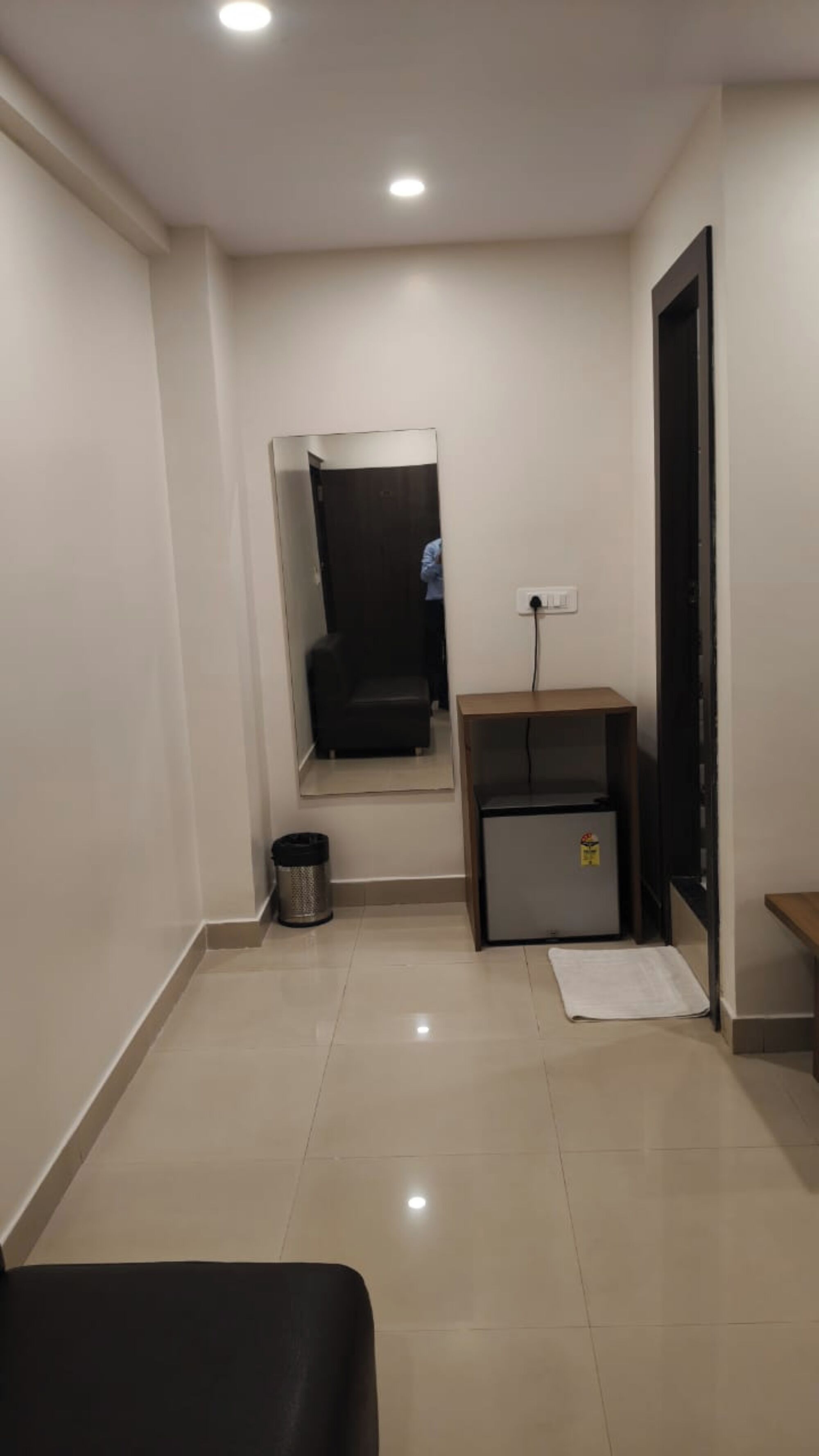 A small, well-lit room with a bed, a desk, a trash bin, and a door.