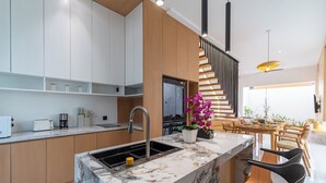 A modern kitchen with a marble island, wooden cabinets, and a dining area with a staircase. - Casa De Vero Amore 3 (Jimbaran)