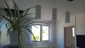 Huoneessa on suuri ruukussa oleva kasvi, ikkunassa verhot, valkoinen patteri, pieni pyöreä pöytä, jonka päällä on koriste-esine, ja ovi, joka johtaa toiseen huoneeseen.