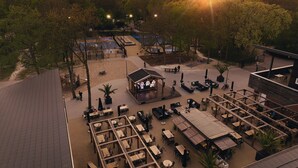 Un área de comedor al aire libre con varias mesas y sillas, rodeada de árboles y un escenario con asientos.