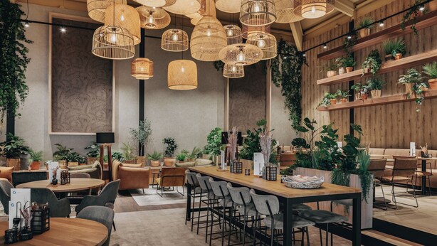 Un interior moderno con varias zonas de estar, incluyendo una mesa comunal larga y varias mesas más pequeñas. Numerosas lámparas de mimbre colgantes ofrecen iluminación ambiental. El espacio está decorado con diversas plantas en macetas, creando un ambiente acogedor.