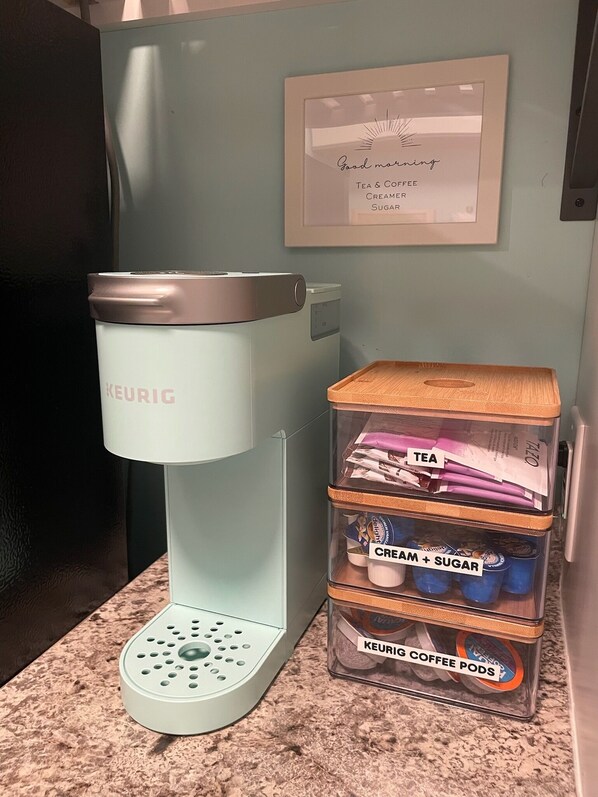 En Keurig-kaffemaskin står på en köksbänk, och bredvid den finns märkta förvaringsbehållare för te, grädde, socker och kaffekapslar.