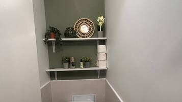 A compact bathroom with a toilet, a shelf with plants and toiletries, and a wall-mounted light fixture.