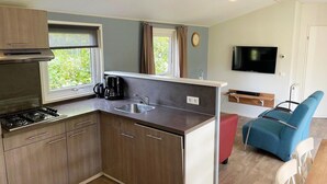 A compact kitchen with a gas stove, a coffee maker, and a sink. There is a TV mounted on the wall, a window with curtains, and a blue chair.