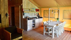 Un hébergement sous tente comprenant une table et des chaises en bois pour les repas, un coin cuisine avec une cuisinière et un four à micro-ondes, et un petit espace salon.