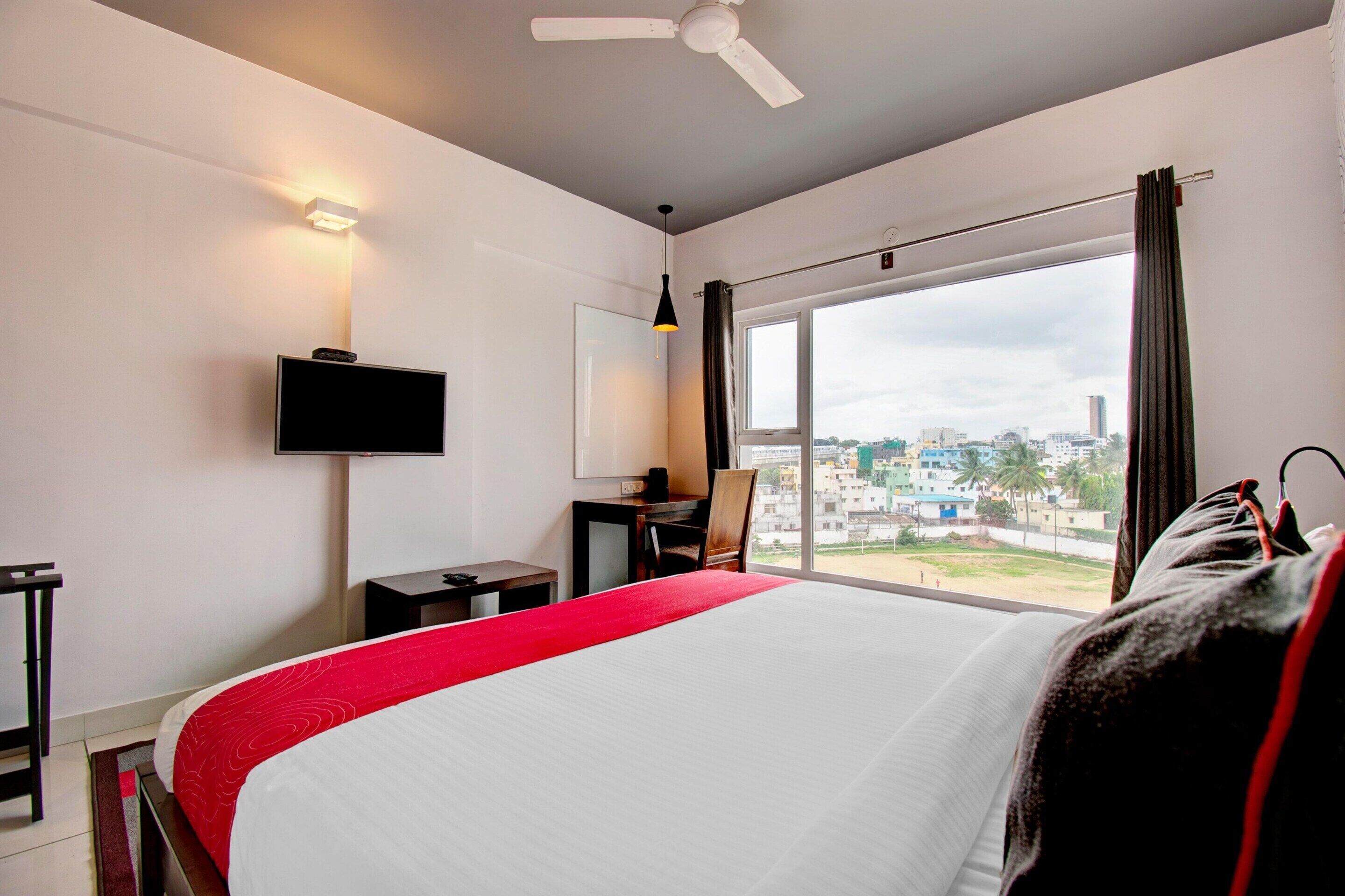 Ein Hotelzimmer mit einem großen Bett, einem Schreibtisch, einem Sessel, einem Fernseher und einem Fenster mit Blick auf die Stadt.