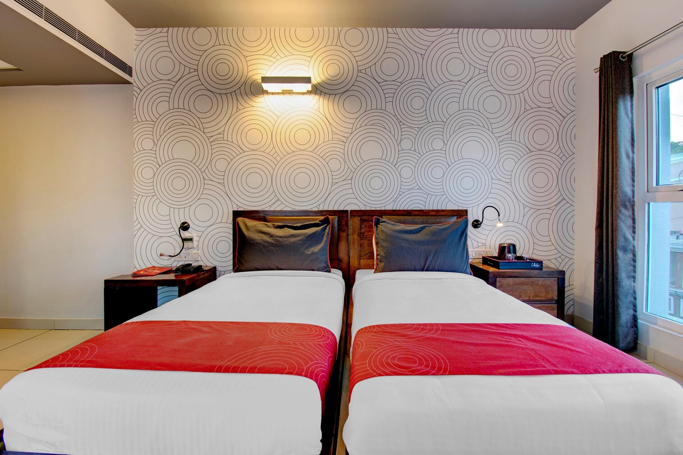 Habitación de hotel con dos camas, papel tapiz estampado, una mesita de noche con lámpara y una ventana con cortinas.