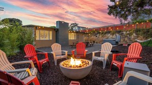 A fire pit surrounded by Adirondack chairs in an outdoor patio area with a view of a building and a hillside. - La Jolla Oasis | 4BR Luxury | Hot Tub | Close to Beach | Sleeps 12 (San Diego)