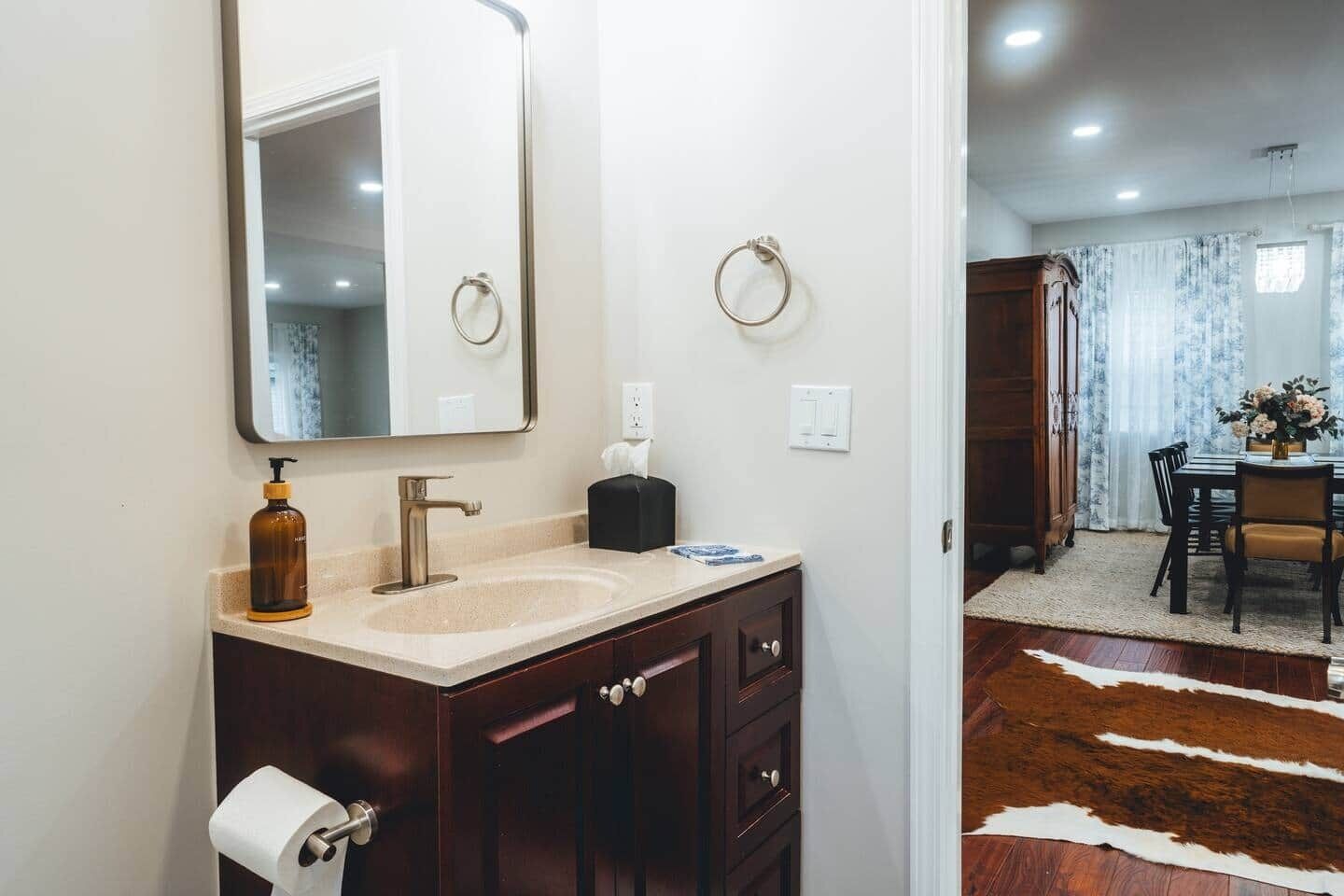 A bathroom with a sink, mirror, and toiletries. There is a dining area with a table and chairs visible in the background.