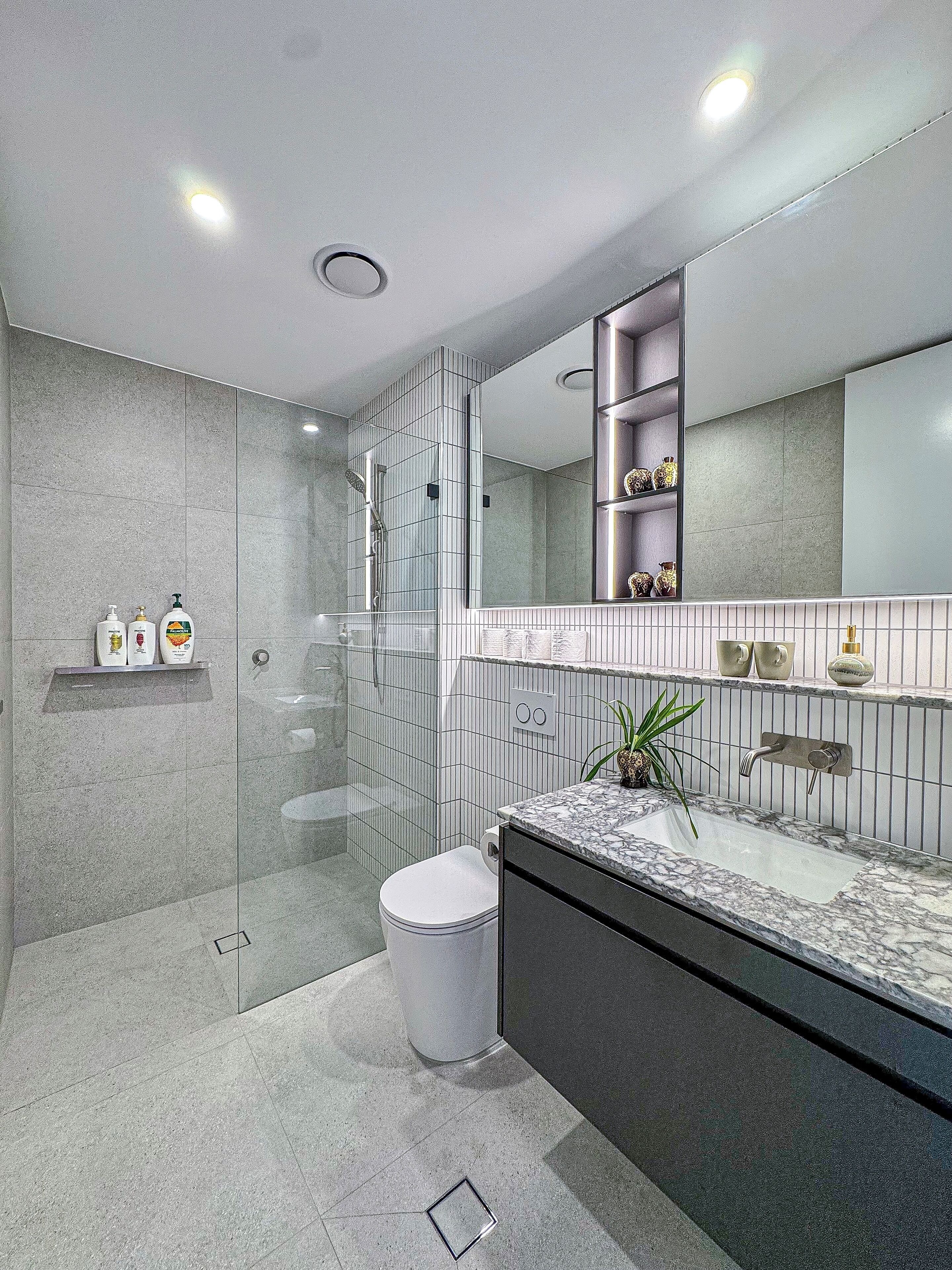 A modern bathroom with a glass shower enclosure, a white toilet, a marble sink, and a large mirror with shelves above.