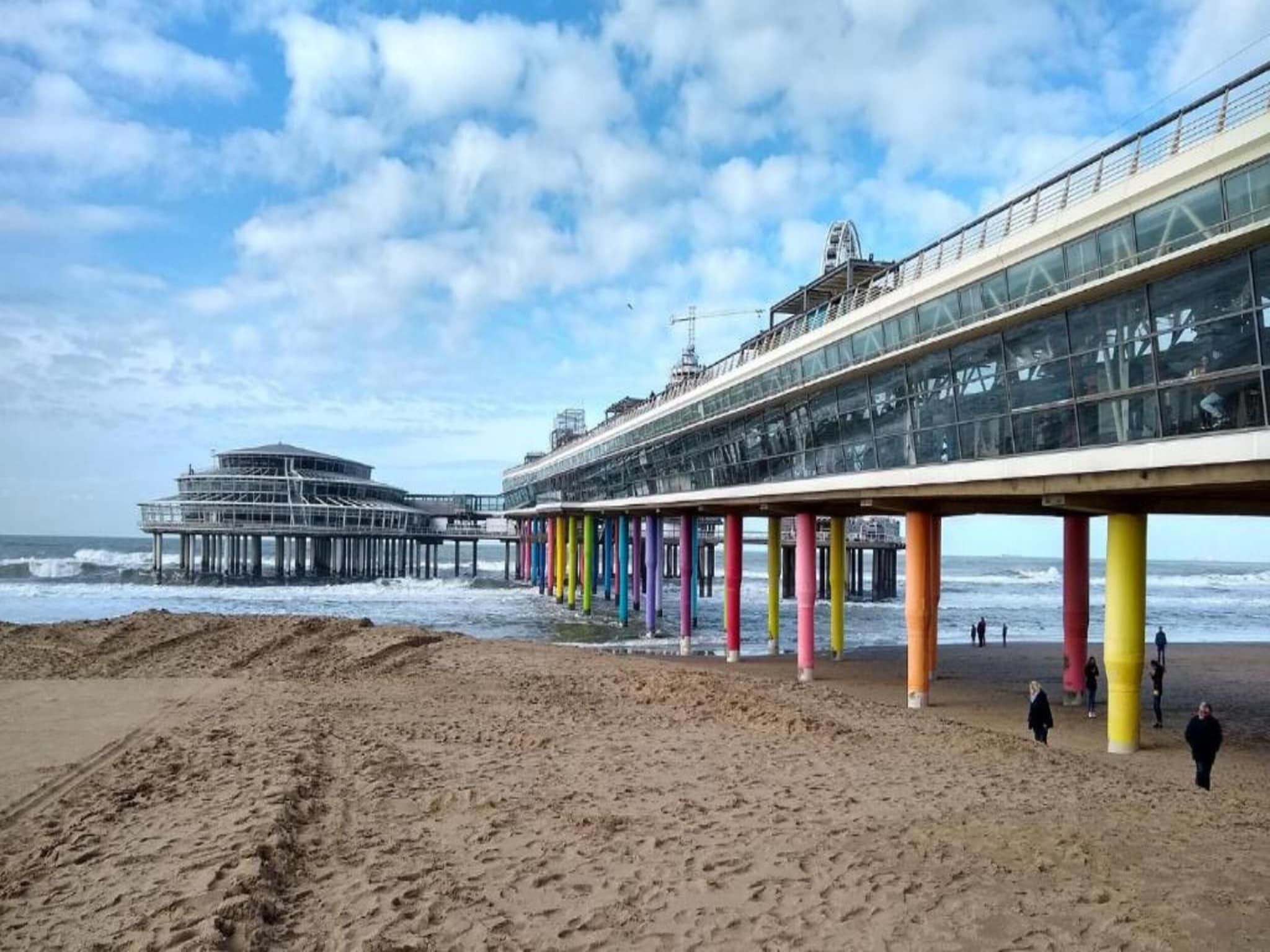 Un muelle con pilares coloridos que se adentra en el mar, un edificio grande con fachada de vidrio y una playa de arena.