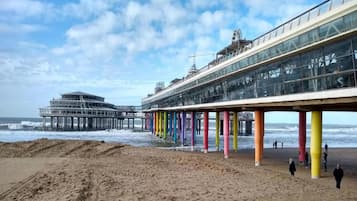 Un muelle con pilares coloridos que se adentra en el mar, un edificio grande con fachada de vidrio y una playa de arena.