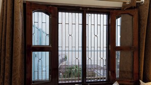 A room with a large window featuring metal bars, a wooden door, and a view of a building outside. - Varanasi Rest House  (Varanasi)