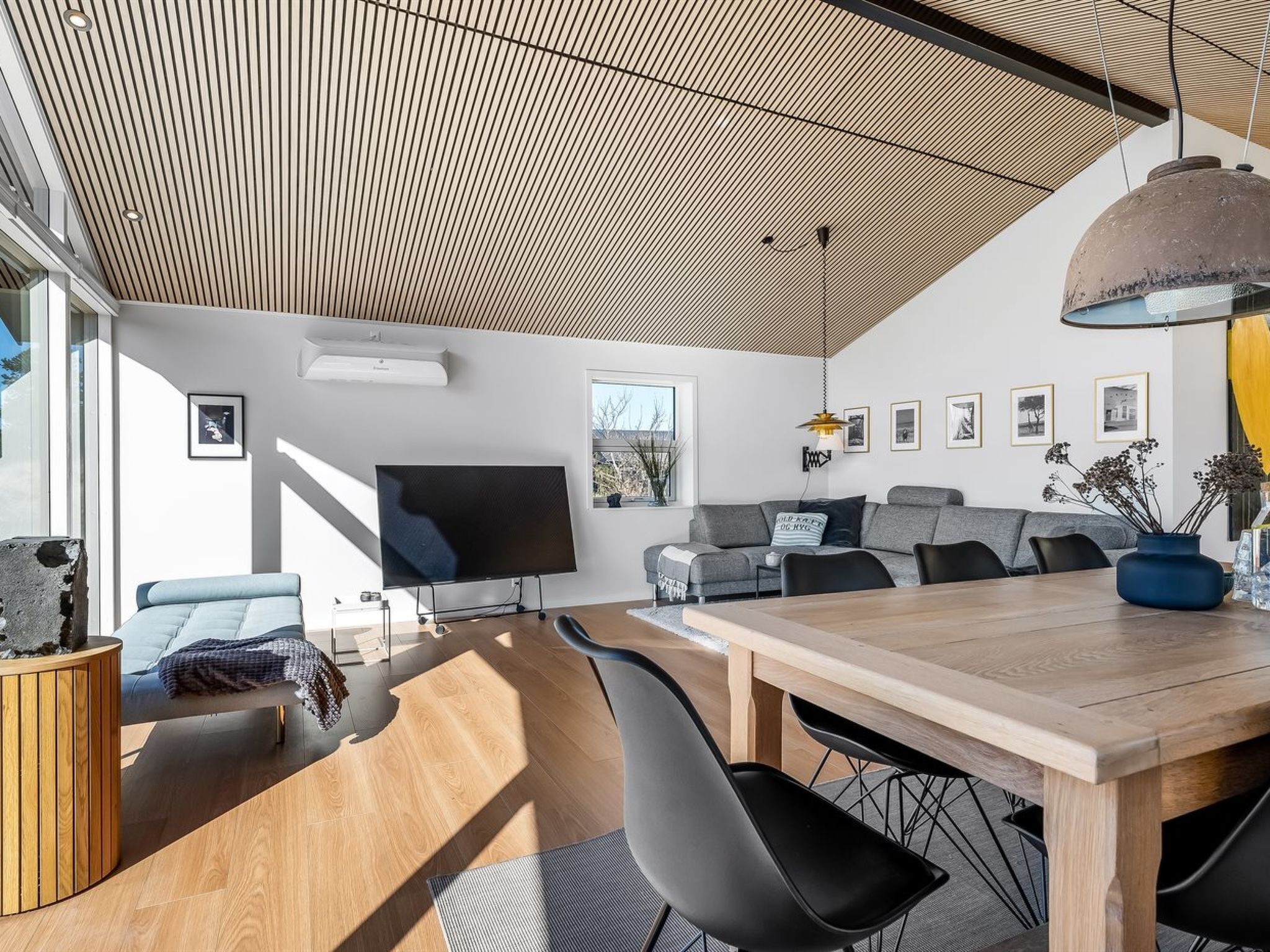 Een moderne woonkamer met een houten plafond, een flatscreen televisie, grijze banken, een eettafel met stoelen en een groot raam.