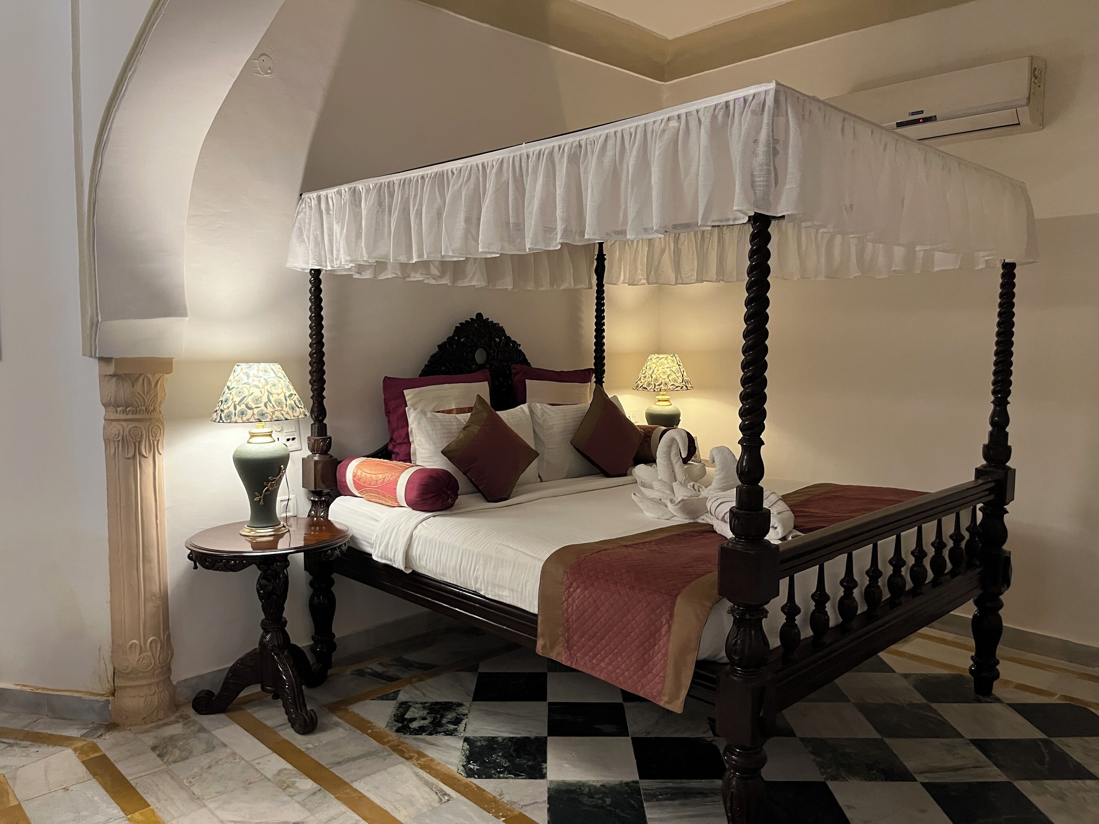A four-poster canopy bed with a checkered floor, two bedside tables with lamps, and a decorative headboard.