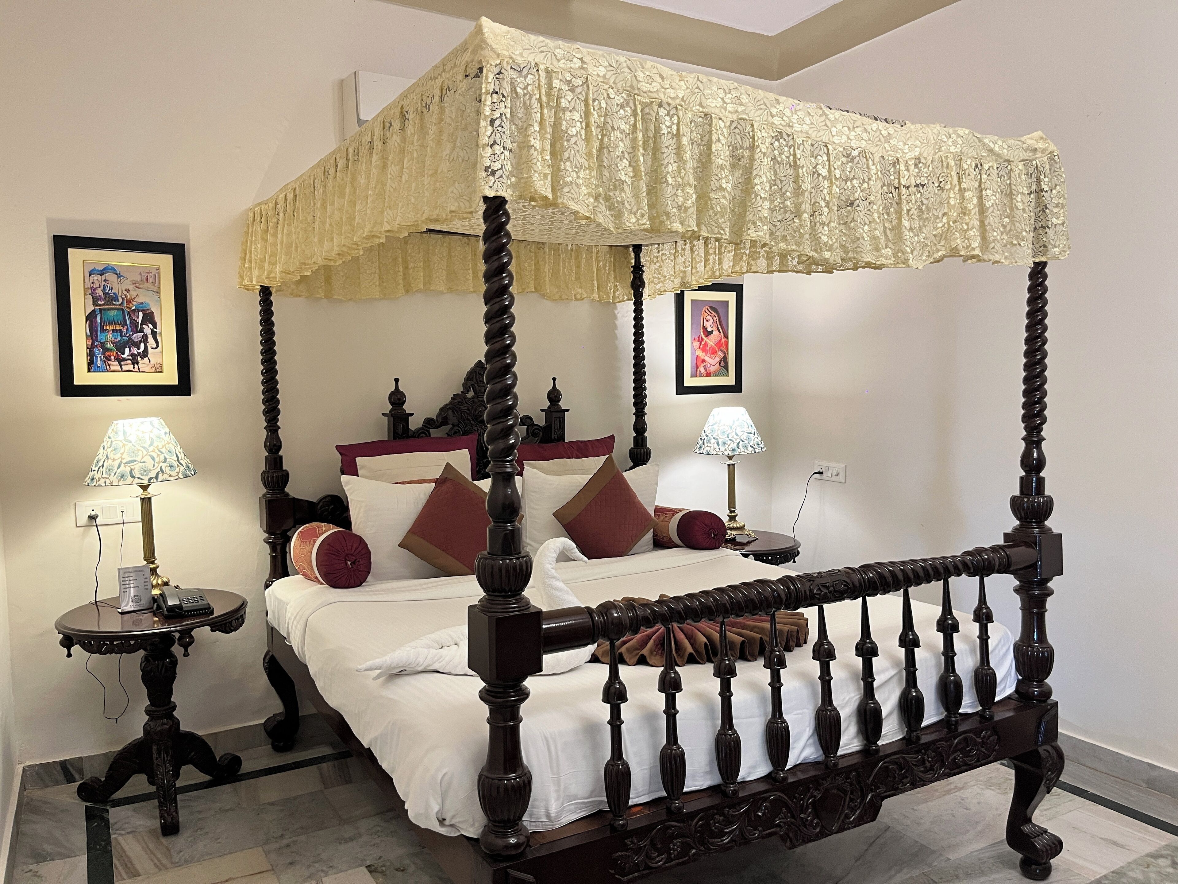A four-poster canopy bed with a white bedspread and multiple pillows, flanked by two bedside tables with lamps, in a room with framed artwork on the walls.