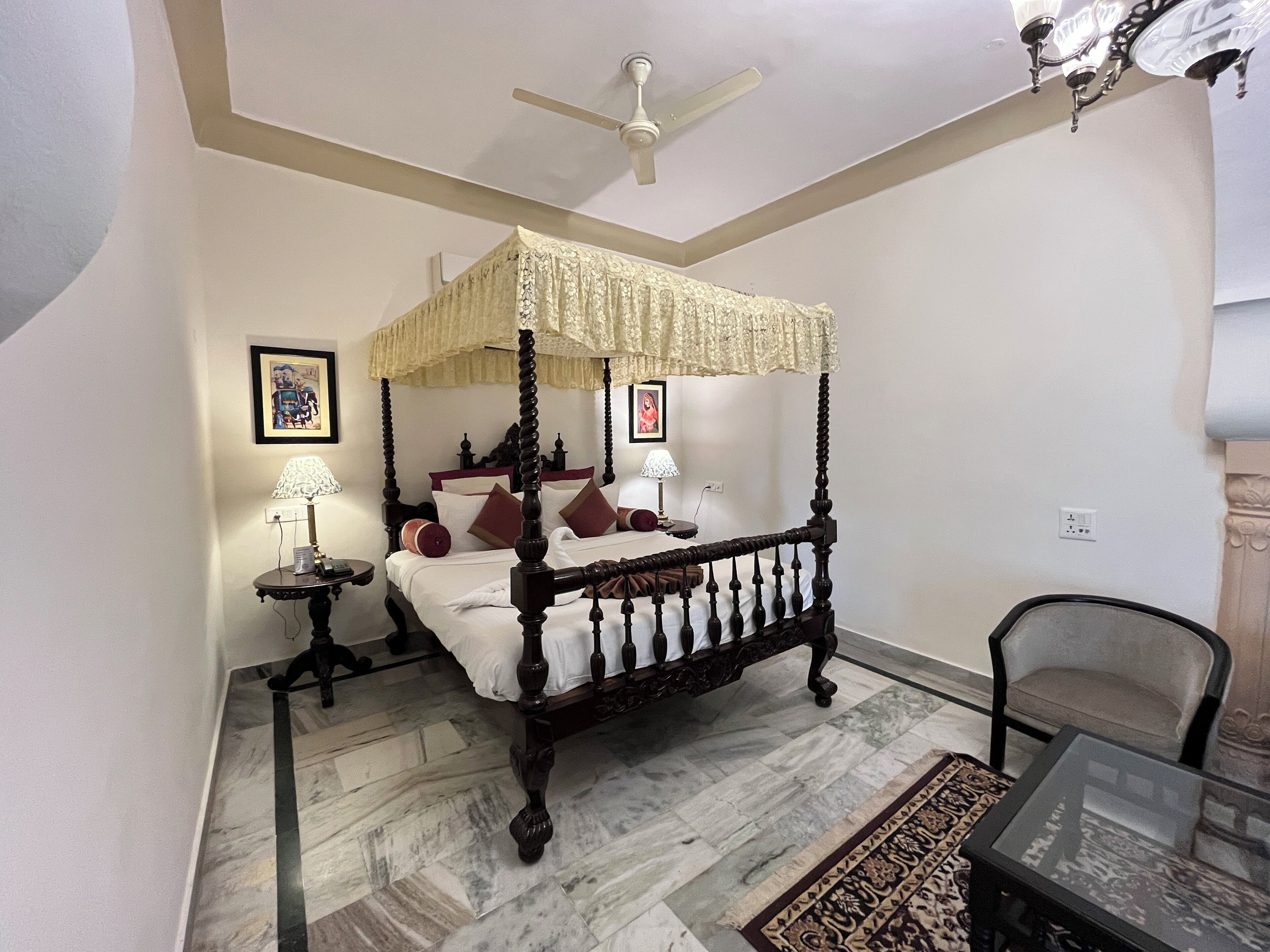 A four-poster canopy bed with a ceiling fan, a side table with a lamp, and a chair in a room with patterned flooring.