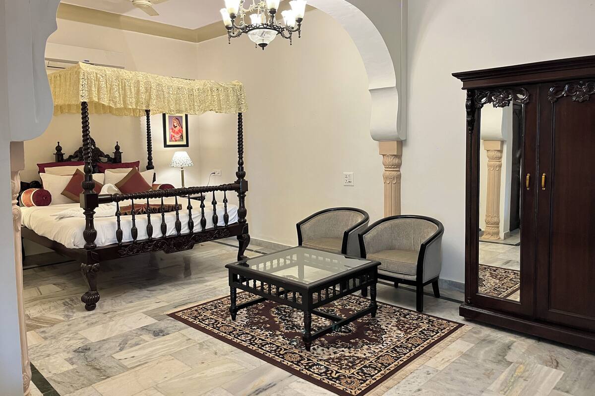 A bedroom with a four-poster bed, a glass-top table, two chairs, a large arched doorway, and a dark wooden wardrobe.