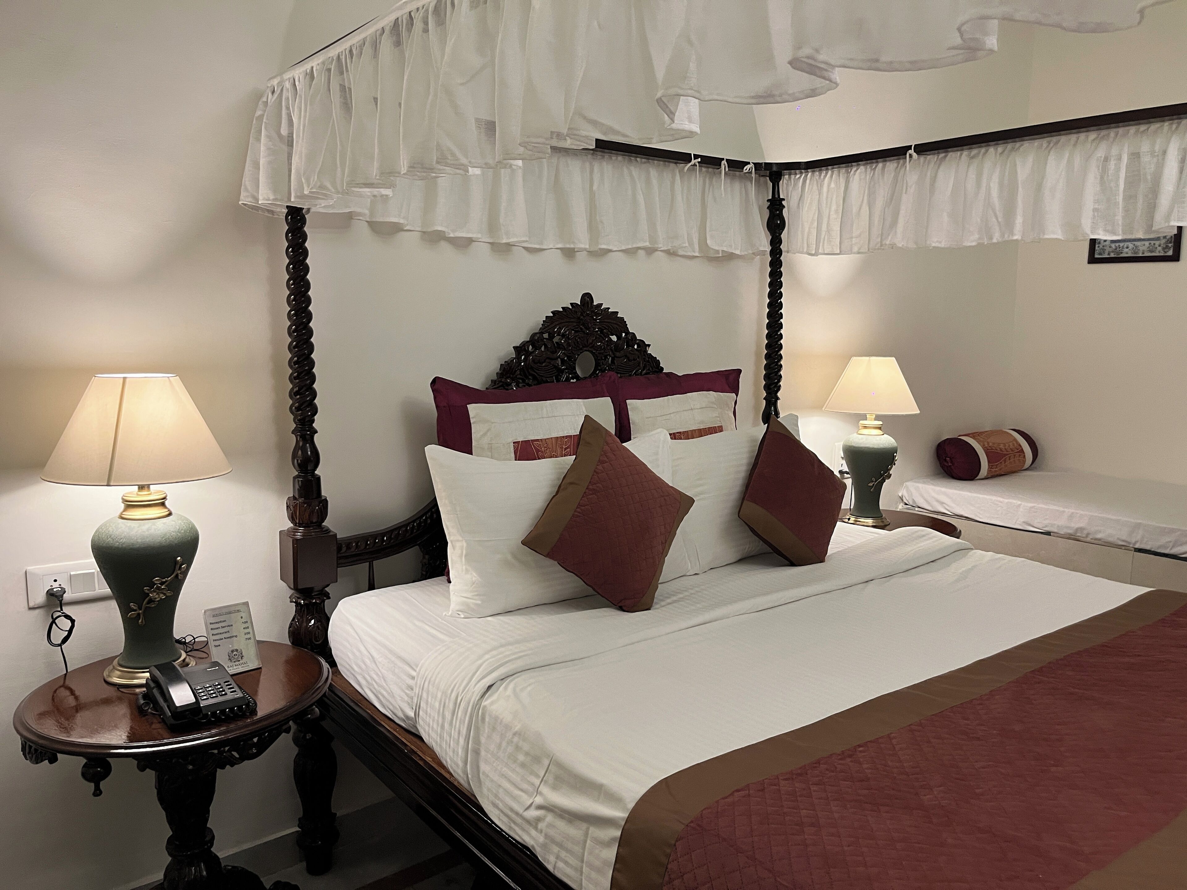 A four-poster bed with white bedding and brown pillows, two bedside tables with lamps, and a canopy overhead.