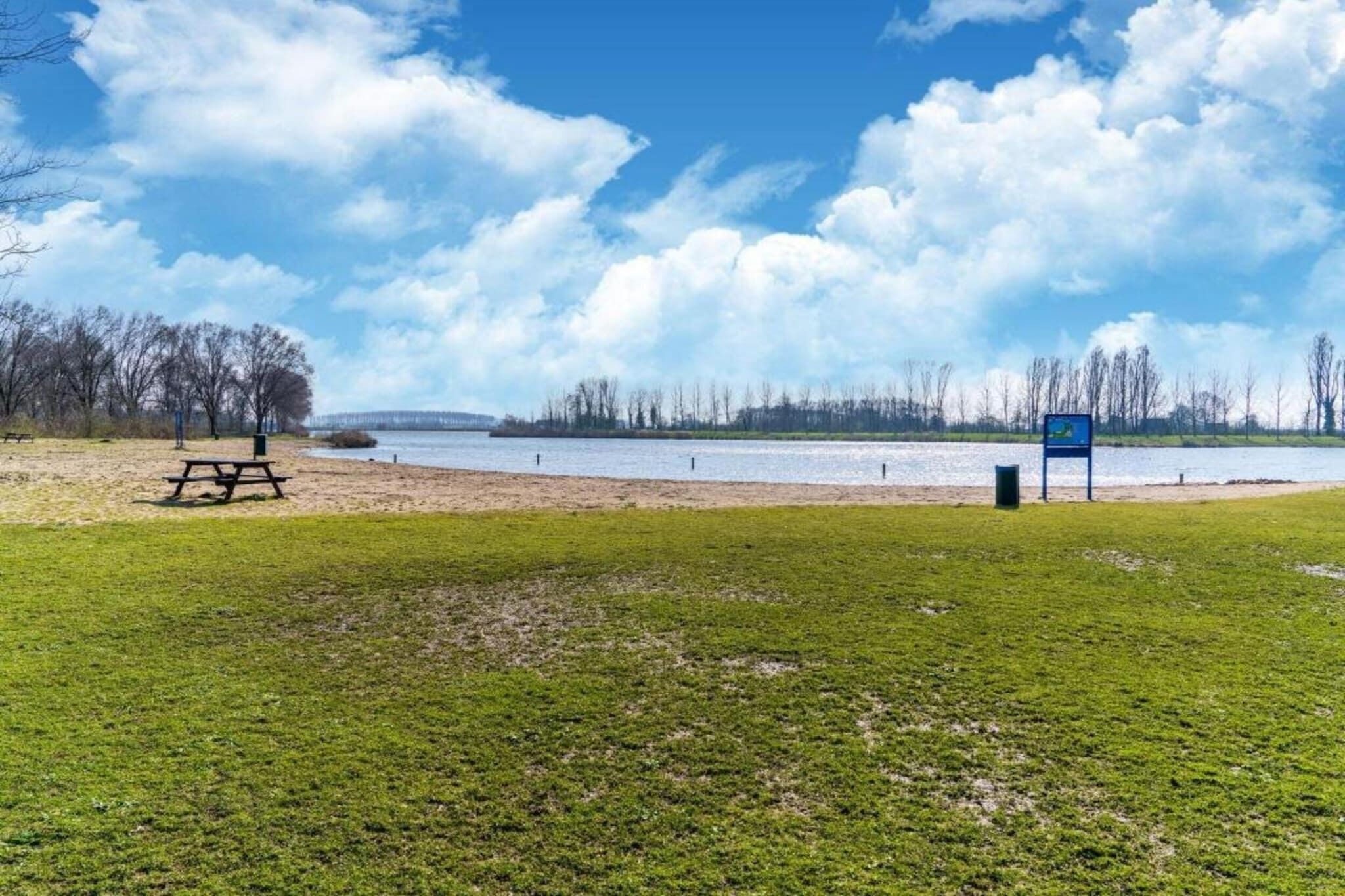 A lakeside park with picnic tables, a trash bin, and a blue sign.