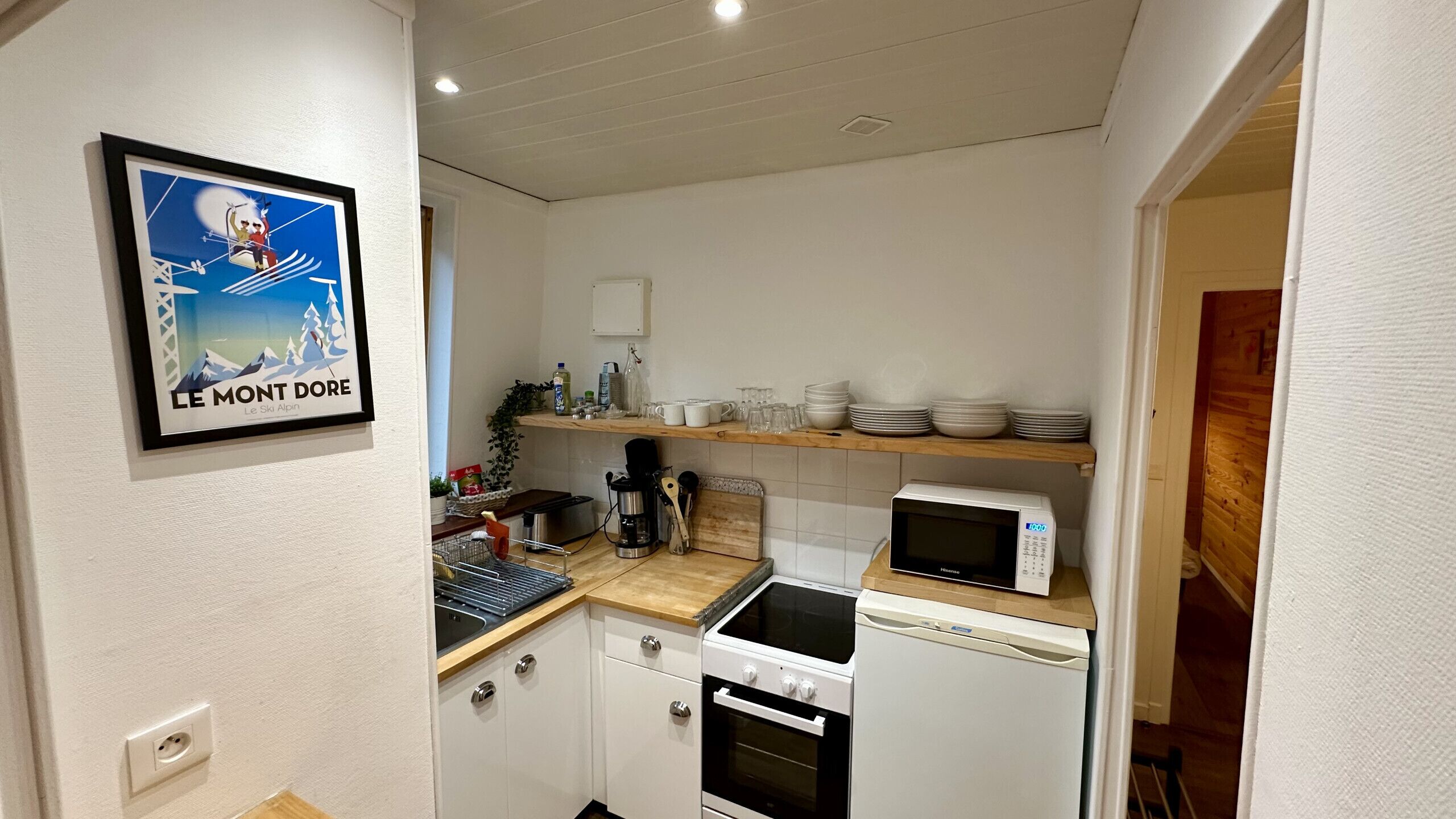 A compact kitchen with white cabinets, a microwave, a stove, and a sink. There is a framed picture on the wall and a shelf with dishes and glasses.