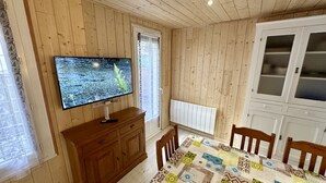 A room with wooden panel walls, a television mounted on the wall, a wooden cabinet, a dining table with a patterned tablecloth, and a white cabinet with glass doors.