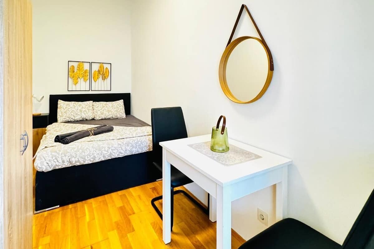 Een kleine, goed verlichte kamer met een bed, een tafel en een stoel. Er hangt een spiegel aan de muur en twee ingekaderde foto’s.
