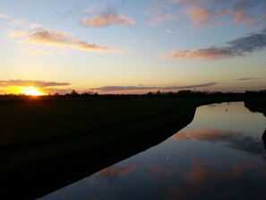 Un coucher de soleil sur une étendue d’eau calme, avec un champ herbeux et une ligne d’arbres à l’horizon.