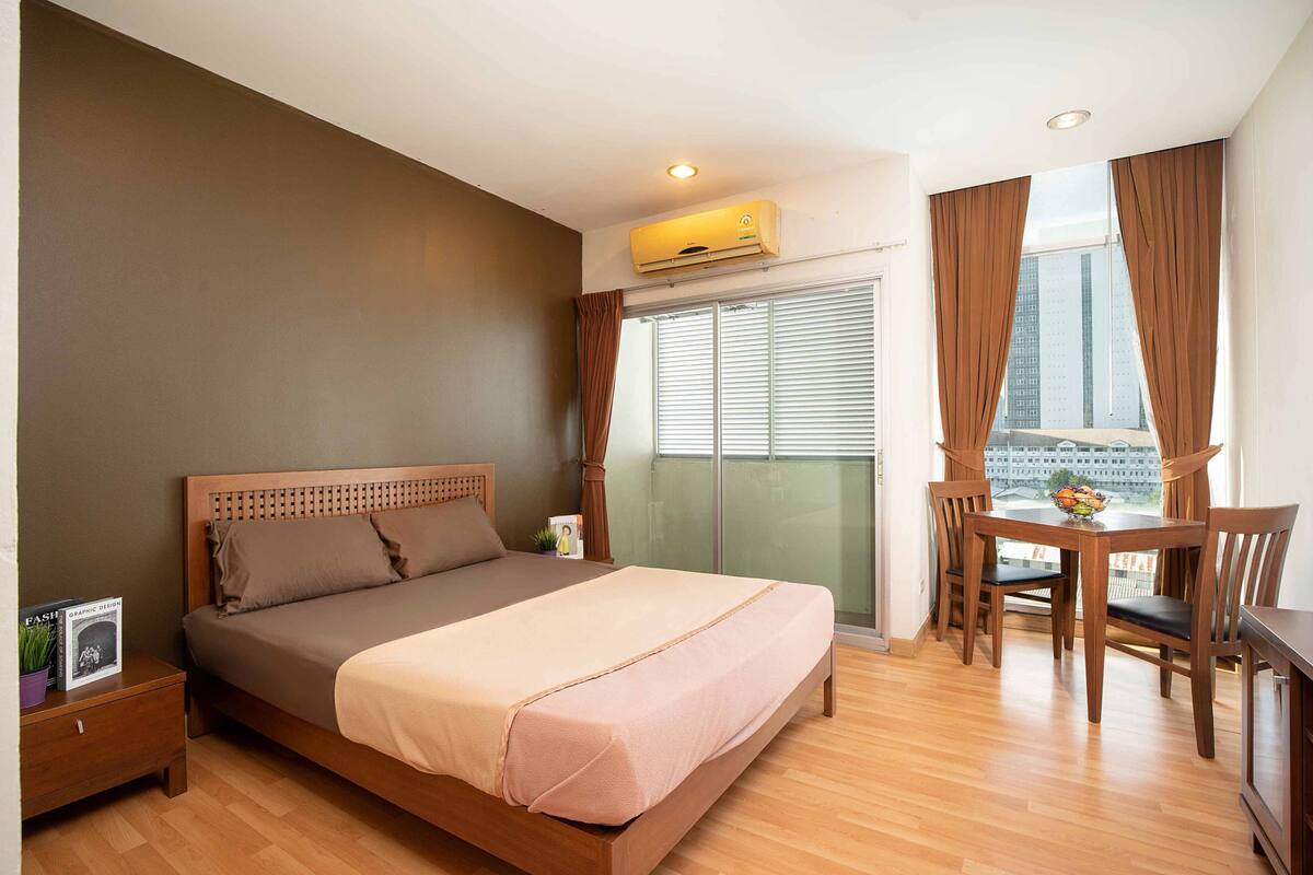 A bedroom with a bed, a nightstand, a dining table with chairs, and a view of a cityscape through the window.
