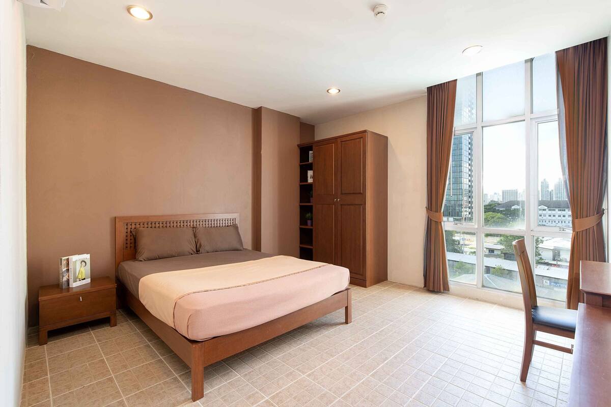 A bedroom with a bed, nightstand, wardrobe, and a view of the city through large windows.